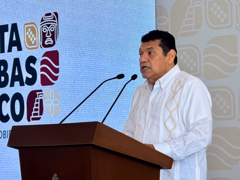Otografía En Plano Medio Del Gobernador Javier May Rodríguez De Pie En Un Podio, Hablando Por Dos Micrófonos. Lleva Una Guayabera Blanca Bordada. Detrás De Él, Una Gran Pantalla Azul Muestra Logotipos De Estilo Gráfico Prehispánico En Blanco, Marrón Y Naranja, Y Parte De La Palabra &Amp;Quot;Tabasco&Amp;Quot; Es Visible. Javier May Mira Hacia La Derecha Mientras Habla.