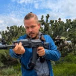 El Actor Tabasqueño Oswaldo Álvarez Caracterizado Con Ropa Casual Y Sosteniendo Un Arma Larga En Un Entorno De Campo Con Nopales, Durante Su Participación En Una Producción Dramática.