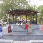 Una Fotografía De Una Plaza De Parque Vibrante Donde Adultos Mayores Bailan Animadamente. La Escena Conserva La Composición De La Imagen Original, Con Las Parejas Y Los Músicos En El Quiosco, Pero Se Han Añadido Elementos Que Celebran La &Quot;Cultura&Quot;. En Primer Plano A La Izquierda, Hay Un Elegante Poste De Hierro Con Un Pergamino Que Dice &Quot;Nuestra Cultura Está Viva: Celebrando El Día Del Abuelo&Quot;. La Base Del Quiosco Tiene Un Cartel De Latón Que Dice &Quot;Ritmos De Nuestra Cultura&Quot;. Un Estandarte Entre Los Árboles Detrás Dice &Quot;Cultura Y Tradición&Quot;. La Iluminación Sigue Siendo Cálida Y Acogedora.
