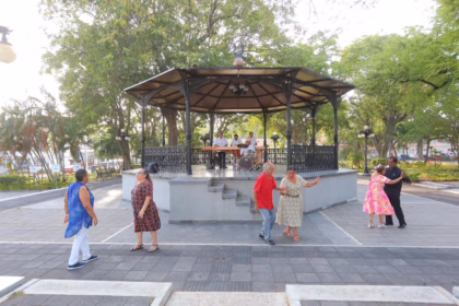 Una Fotografía De Una Plaza De Parque Vibrante Donde Adultos Mayores Bailan Animadamente. La Escena Conserva La Composición De La Imagen Original, Con Las Parejas Y Los Músicos En El Quiosco, Pero Se Han Añadido Elementos Que Celebran La &Quot;Cultura&Quot;. En Primer Plano A La Izquierda, Hay Un Elegante Poste De Hierro Con Un Pergamino Que Dice &Quot;Nuestra Cultura Está Viva: Celebrando El Día Del Abuelo&Quot;. La Base Del Quiosco Tiene Un Cartel De Latón Que Dice &Quot;Ritmos De Nuestra Cultura&Quot;. Un Estandarte Entre Los Árboles Detrás Dice &Quot;Cultura Y Tradición&Quot;. La Iluminación Sigue Siendo Cálida Y Acogedora.