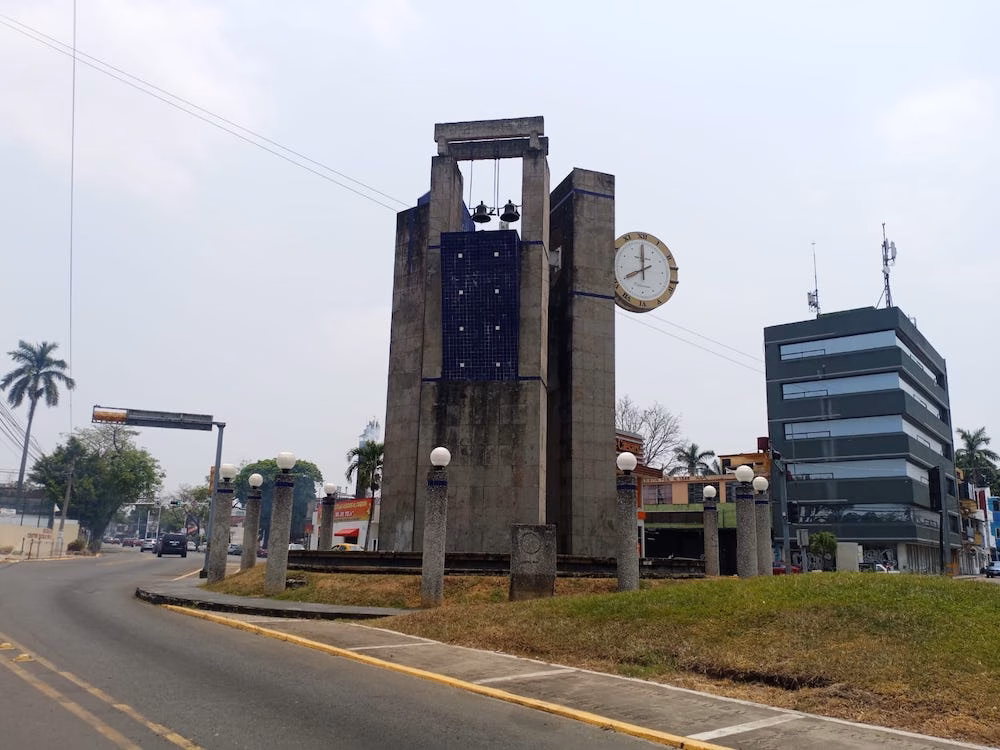 ¿Y Tú, Ya Lo Conoces? La Historia, Arquitectura E Identidad Detrás Del Reloj De Las Tres Caras 73 Vista Panorámica Del Monumento Del Reloj De Las Tres Caras En La Glorieta De Paseo De La Sierra, Mostrando Su Estructura De Concreto Con Campanas Superiores, Carátula Circular Y Entorno Urbano.