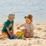 Dos Niños Con Sombreros Para El Sol Jugando Con Balde Y Pala En La Arena A La Orilla Del Agua, Bajo Supervisión En Un Entorno Acuático Seguro.