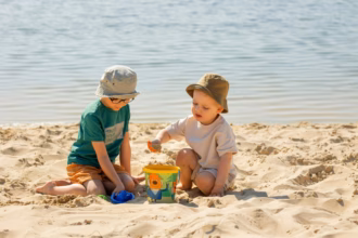 Dos Niños Con Sombreros Para El Sol Jugando Con Balde Y Pala En La Arena A La Orilla Del Agua, Bajo Supervisión En Un Entorno Acuático Seguro.