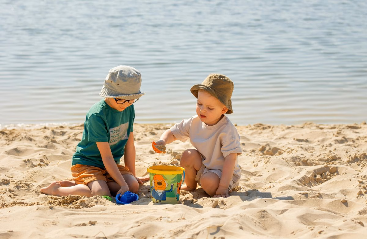 Verano Seguro: Recomendaciones Clave Para Disfrutar De Balnearios Y Ríos En Tabasco 70 Dos Niños Con Sombreros Para El Sol Jugando Con Balde Y Pala En La Arena A La Orilla Del Agua, Bajo Supervisión En Un Entorno Acuático Seguro.