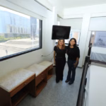 Dos Estudiantes Universitarias Posan En El Interior De Una Habitación Amueblada De La Villa Universitaria En Villahermosa, Con Literas, Muebles De Madera Y Una Gran Ventana Con Vista Al Exterior.