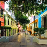 Ista A Nivel De Calle De Un Pasaje Peatonal Dentro De La Zona Luz, Caracterizado Por Edificios Coloniales Pintados Con Colores Vivos (Rosa, Verde, Naranja Y Azul), Árboles Que Dan Sombra Al Camino Y Luminarias Clásicas Al Atardecer.