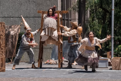 Uatro Actores Actúan En Una Obra De Teatro Callejero En Un Parque. Un Actor Lleva Una Estructura De Madera Con Un Panel De Madera, Una Actriz Lleva Un Disfraz De Mujer Gorda, Un Actor Lleva Un Disfraz De Granjero Y Una Actriz Lleva Un Vestido Con Capas Y Tiene Los Brazos Extendidos. Hay Público Sentado Detrás De Ellos.