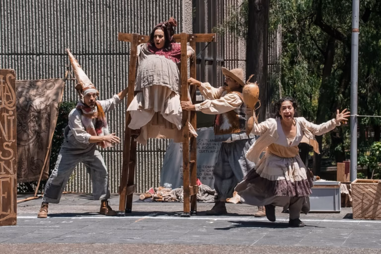 Uatro Actores Actúan En Una Obra De Teatro Callejero En Un Parque. Un Actor Lleva Una Estructura De Madera Con Un Panel De Madera, Una Actriz Lleva Un Disfraz De Mujer Gorda, Un Actor Lleva Un Disfraz De Granjero Y Una Actriz Lleva Un Vestido Con Capas Y Tiene Los Brazos Extendidos. Hay Público Sentado Detrás De Ellos.