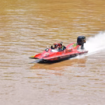 &Quot;Una Lancha De Carreras Roja Con El Número 358 Navega A Gran Velocidad Por El Agua Marrón De Un Río, Creando Una Estela De Agua Blanca A Su Paso. Dos Personas Con Cascos Están A Bordo.&Quot;