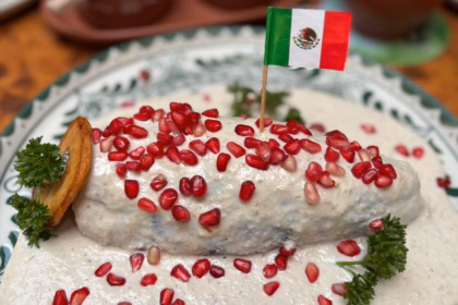 Chile En Nogada Tradicional Servido Sobre Un Plato Decorado, Cubierto Con Crema De Nuez De Castilla, Granos De Granada Roja, Perejil Y Una Pequeña Bandera De México.