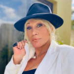 La Cantante Y Actriz Tabasqueña Laura León Posando Al Aire Libre Con Un Sombrero Negro, Saco Blanco Y Un Collar Esférico Metálico.