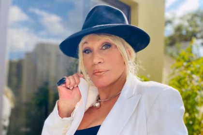 La Cantante Y Actriz Tabasqueña Laura León Posando Al Aire Libre Con Un Sombrero Negro, Saco Blanco Y Un Collar Esférico Metálico.