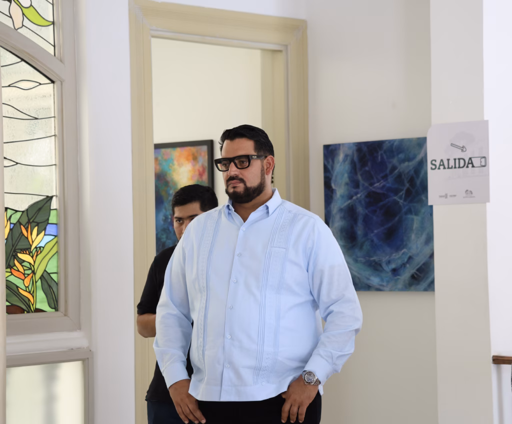 Hospital Zumaya Integrará El Arte Y La Cultura De Tabasco En Sus Pasillos: Alejandro Yusseff 3 El Ceo Del Hospital Zumaya, Alejandro Yusseff Nieves, Posa De Pie En Una Habitación De Estilo Elegante. Viste Guayabera Azul Claro, Anteojos Y Barba. A Su Espalda Se Observa Un Gran Mueble De Madera Oscura Con Compartimentos Y Frente A Él Se Muestra Una Exhibición De Coloridos Vestidos Tradicionales Sobre Maniquíes De Madera, Delimitados Por Postes Dorados Y Cordones Rojos De Protección, Junto A Pequeñas Esculturas Sobre Una Mesa.