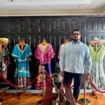Un Hombre Con Barba, Anteojos Y Guayabera Azul Claro (Alejandro Yusseff) Está De Pie En Una Habitación De Estilo Elegante. A Su Espalda Se Observa Un Gran Mueble O Pared De Madera Oscura Con Compartimentos. Frente A Él Hay Una Exhibición Que Incluye Varios Trajes Tradicionales Coloridos Y Ornamentados Colocados En Maniquíes De Madera, Delimitados Por Postes Dorados Y Cordones Rojos De Protección. En Primer Plano Se Aprecia Una Mesa De Madera Con Pequeñas Esculturas Decorativas.