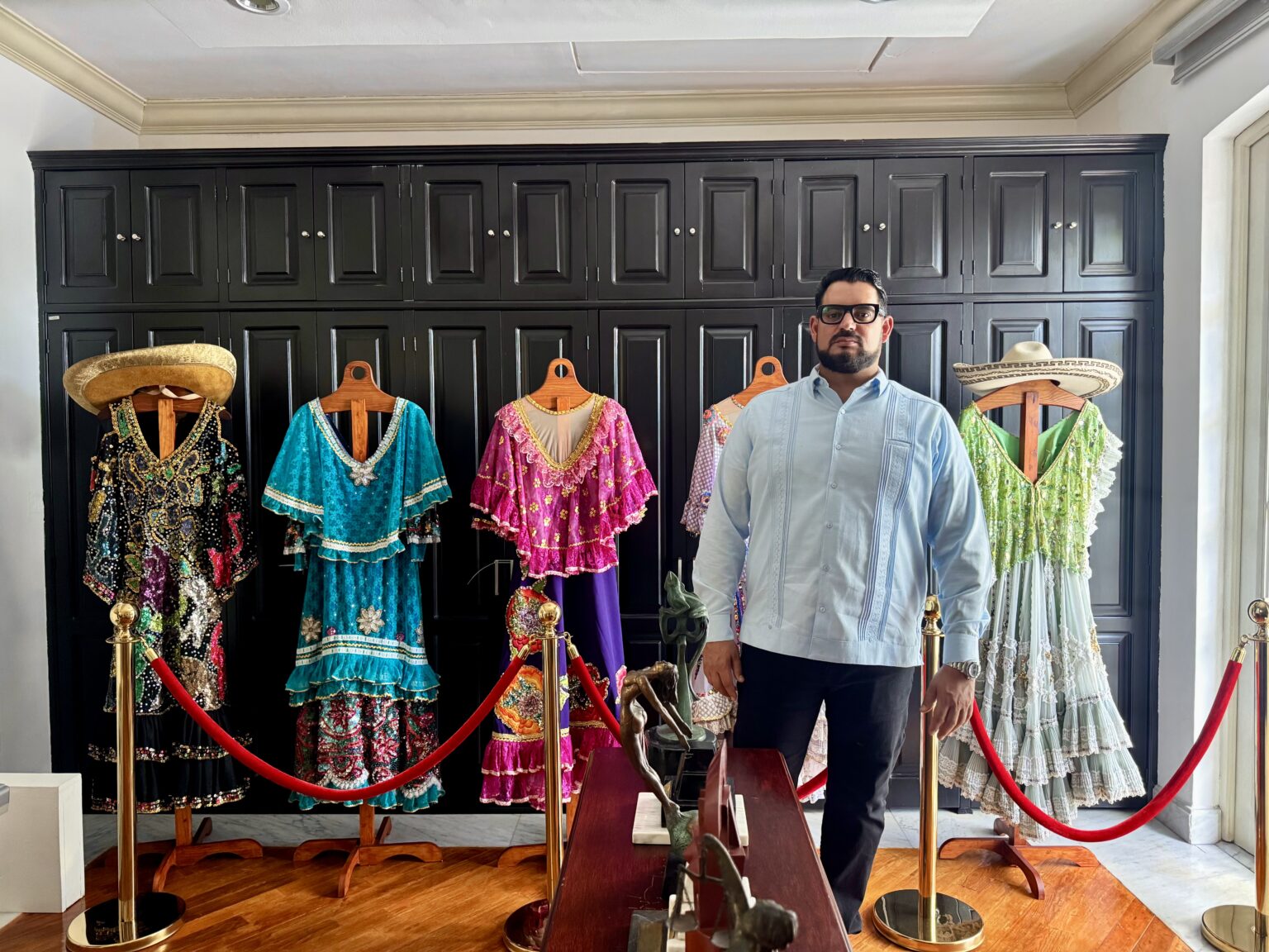 Hospital Zumaya Integrará El Arte Y La Cultura De Tabasco En Sus Pasillos: Alejandro Yusseff 71 Un Hombre Con Barba, Anteojos Y Guayabera Azul Claro (Alejandro Yusseff) Está De Pie En Una Habitación De Estilo Elegante. A Su Espalda Se Observa Un Gran Mueble O Pared De Madera Oscura Con Compartimentos. Frente A Él Hay Una Exhibición Que Incluye Varios Trajes Tradicionales Coloridos Y Ornamentados Colocados En Maniquíes De Madera, Delimitados Por Postes Dorados Y Cordones Rojos De Protección. En Primer Plano Se Aprecia Una Mesa De Madera Con Pequeñas Esculturas Decorativas.