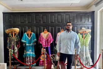 Un Hombre Con Barba, Anteojos Y Guayabera Azul Claro (Alejandro Yusseff) Está De Pie En Una Habitación De Estilo Elegante. A Su Espalda Se Observa Un Gran Mueble O Pared De Madera Oscura Con Compartimentos. Frente A Él Hay Una Exhibición Que Incluye Varios Trajes Tradicionales Coloridos Y Ornamentados Colocados En Maniquíes De Madera, Delimitados Por Postes Dorados Y Cordones Rojos De Protección. En Primer Plano Se Aprecia Una Mesa De Madera Con Pequeñas Esculturas Decorativas.