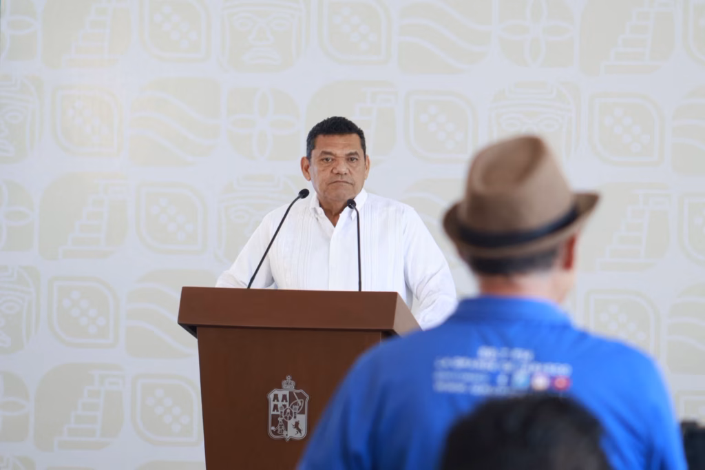 Javier May Presenta Intensa Agenda Semanal: Obras, Programas Sociales, Balance Ambiental Y Avances Legislativos En Tabasco 73 Fotografía Del Gobernador Javier May Rodríguez De Pie Tras Un Podio De Madera Hablando Durante Una Conferencia Matutina En Tabasco. Viste Una Guayabera Blanca Y De Fondo Hay Una Pared Con Logotipos Gubernamentales Estampados. En Primer Plano, De Espaldas, Se Ve Parcialmente A Un Asistente Con Camisa Azul Y Sombrero Marrón.