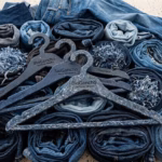 Tres Perchas De Ropa Sostenibles, De Color Azul Con Motas Blancas Y Grabadas Con La Inscripción &Quot;Biogancho - Hecho En Tabasco - Reciclado&Quot;, Están Dispuestas En Primer Plano Sobre Una Colección De Vaqueros Enrollados Y Restos De Tela Vaquera Deshilachada Sobre Una Superficie Jaspeada.