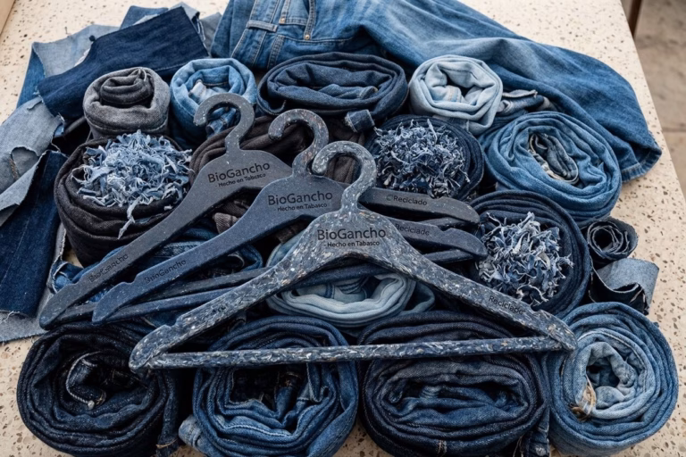 Tres Perchas De Ropa Sostenibles, De Color Azul Con Motas Blancas Y Grabadas Con La Inscripción &Amp;Quot;Biogancho - Hecho En Tabasco - Reciclado&Amp;Quot;, Están Dispuestas En Primer Plano Sobre Una Colección De Vaqueros Enrollados Y Restos De Tela Vaquera Deshilachada Sobre Una Superficie Jaspeada.