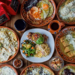 Mesa Servida Con Variados Platillos Tradicionales En Vajilla De Barro, Incluyendo Chilaquiles, Huevos Rancheros, Enchiladas, Ensalada Y Café De Olla Sobre Mesa De Madera.