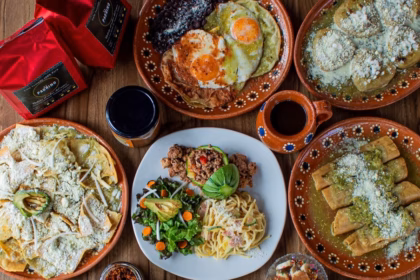 Mesa Servida Con Variados Platillos Tradicionales En Vajilla De Barro, Incluyendo Chilaquiles, Huevos Rancheros, Enchiladas, Ensalada Y Café De Olla Sobre Mesa De Madera.