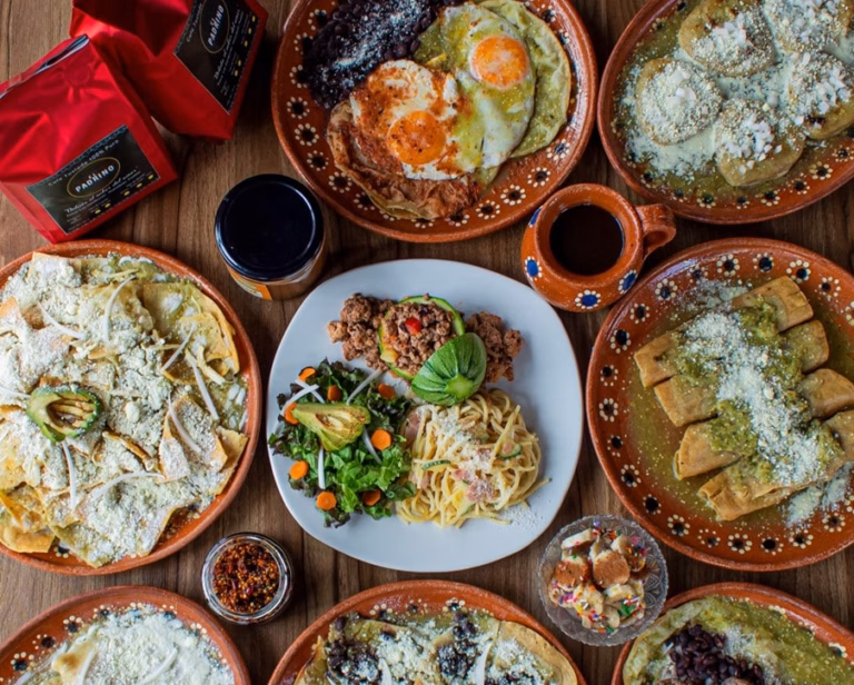 Mesa Servida Con Variados Platillos Tradicionales En Vajilla De Barro, Incluyendo Chilaquiles, Huevos Rancheros, Enchiladas, Ensalada Y Café De Olla Sobre Mesa De Madera.