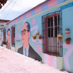 Vista De Ángulo Bajo De Un Vibrante Mural Callejero Que Cubre La Pared De Un Edificio. El Mural Representa Retratos Estilizados De Personajes Históricos Con Gafas, Intercalados Con Motivos Florales Abstractos Y Líneas Geométricas. La Pared Tiene Macetas Colgantes Y Una Ventana Con Rejas. El Suelo Es De Adoquines De Terracota.