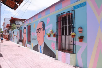 Vista De Ángulo Bajo De Un Vibrante Mural Callejero Que Cubre La Pared De Un Edificio. El Mural Representa Retratos Estilizados De Personajes Históricos Con Gafas, Intercalados Con Motivos Florales Abstractos Y Líneas Geométricas. La Pared Tiene Macetas Colgantes Y Una Ventana Con Rejas. El Suelo Es De Adoquines De Terracota.