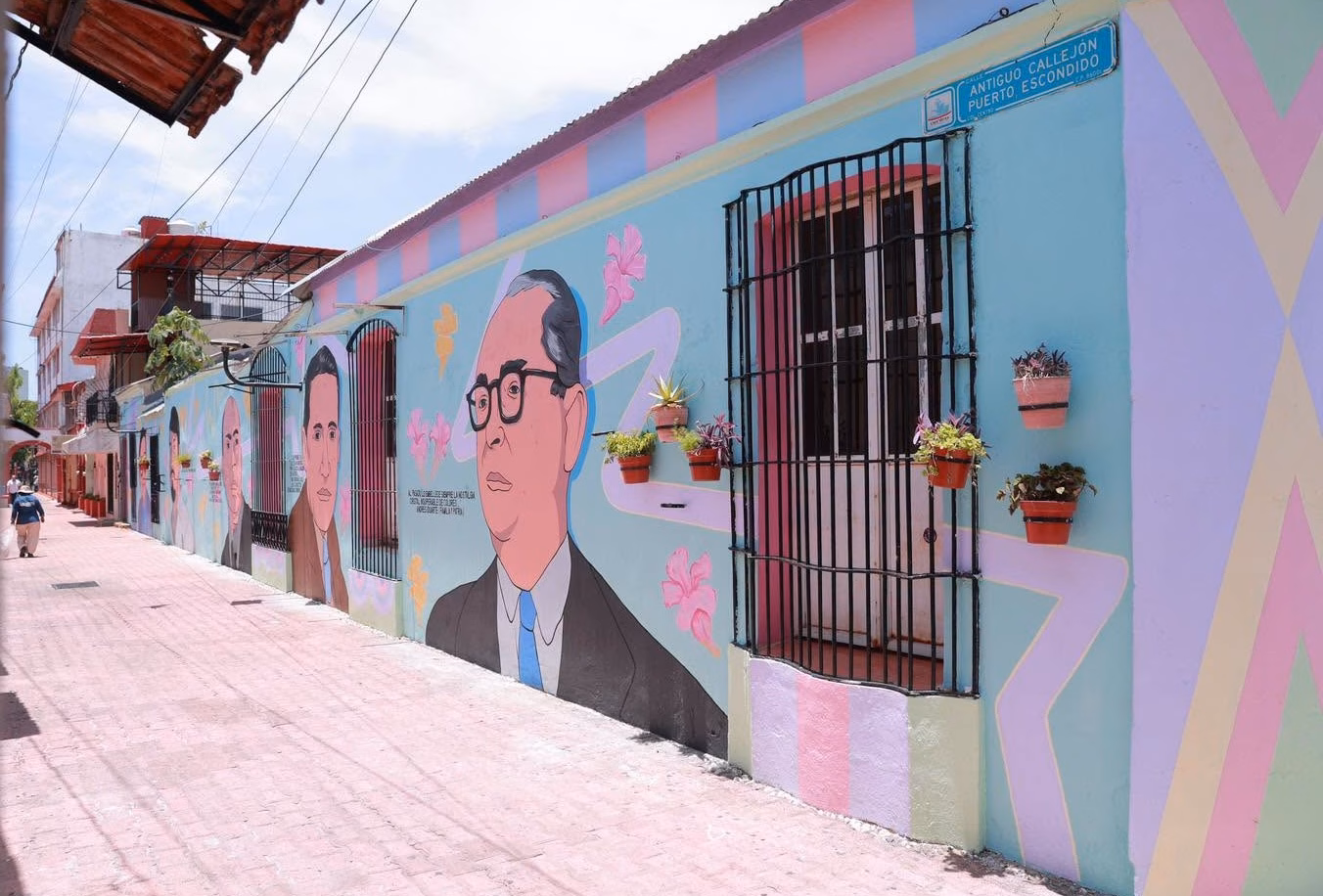 Vista De Ángulo Bajo De Un Vibrante Mural Callejero Que Cubre La Pared De Un Edificio. El Mural Representa Retratos Estilizados De Personajes Históricos Con Gafas, Intercalados Con Motivos Florales Abstractos Y Líneas Geométricas. La Pared Tiene Macetas Colgantes Y Una Ventana Con Rejas. El Suelo Es De Adoquines De Terracota.