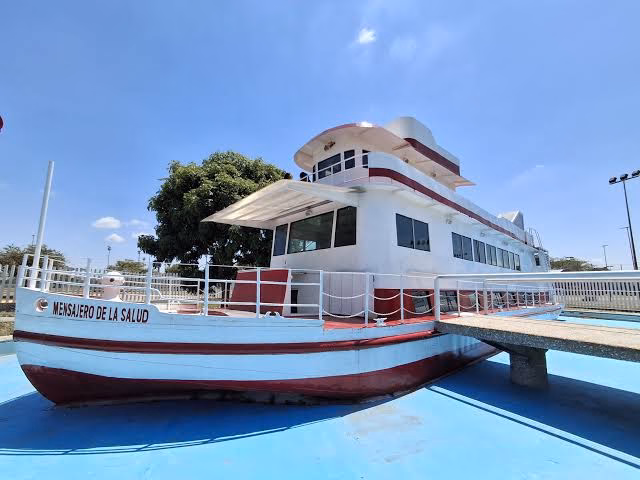 Una Toma De Ángulo Bajo De La Histórica Embarcación &Quot;Mensajero De La Salud&Quot; En Exhibición, Pintada De Blanco Y Rojo Sobre Una Superficie Azul Simulando Agua, Con Un Muelle De Concreto Que Conduce A Ella Bajo Un Cielo Despejado.