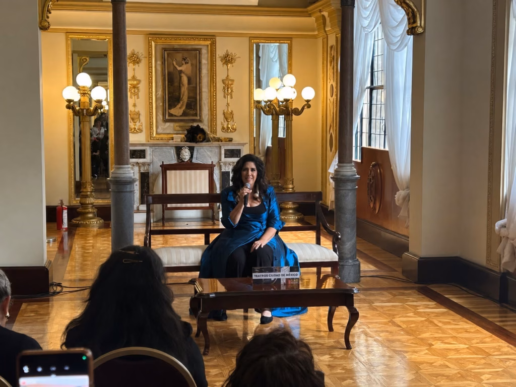 Regina Orozco Recuerda A La Tabasqueña Esperanza Iris Y Revela Historia De Aparecidos En Su Teatro 73 Una Vista Más Amplia De La Lujosa Sala De Estar Con Regina Orozco Sentada En Un Banco Acolchado En El Centro. Regina Lleva El Mismo Abrigo De Satén Azul Y Está Hablando Por Un Micrófono, Mirando Hacia El Público. Un Pequeño Cartel En La Mesa Baja Frente A Ella Dice &Quot;Teatros Ciudad De México&Quot;. La Sala Está Llena Con Un Público Sentado En Primer Plano, Con La Cabeza Y Los Hombros De Varias Personas Visibles. Los Elementos De Diseño De La Sala, Incluyendo Las Columnas Y Los Candelabros Dorados, Son Más Prominentes Desde Este Ángulo.