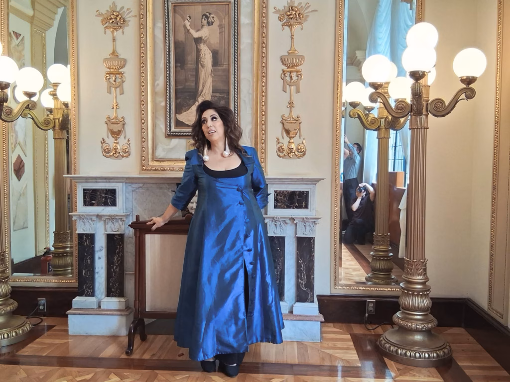 Regina Orozco Recuerda A La Tabasqueña Esperanza Iris Y Revela Historia De Aparecidos En Su Teatro 72 Una Toma De Plano Medio De La Actriz Y Cantante Regina Orozco De Pie En Una Lujosa Sala De Estar De Estilo Palaciego. Regina Lleva Un Abrigo Largo De Satén Azul Eléctrico Sobre Un Atuendo Negro. Sonríe Y Mira Hacia Su Derecha, Con La Mano Izquierda Apoyada Ligeramente Sobre Un Soporte De Madera. La Sala Cuenta Con Intrincadas Molduras Doradas, Una Chimenea De Mármol, Candelabros Dorados Ornamentados Y Pisos De Parquet. Dos Personas Con Cámaras Son Visibles En El Reflejo De Un Gran Espejo A La Derecha.