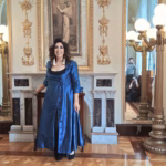 La Soprano Y Actriz Mexicana Regina Orozco Posa Sonriente Con Un Abrigo Largo De Satén Azul En El Histórico Foyer Del Teatro De La Ciudad Esperanza Iris, Luciendo Un Peinado Ondulado Frente A La Chimenea De Mármol Y Retratos De Época.