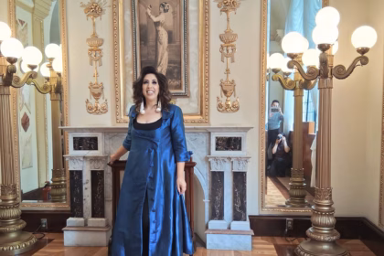 La Soprano Y Actriz Mexicana Regina Orozco Posa Sonriente Con Un Abrigo Largo De Satén Azul En El Histórico Foyer Del Teatro De La Ciudad Esperanza Iris, Luciendo Un Peinado Ondulado Frente A La Chimenea De Mármol Y Retratos De Época.