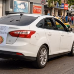 Vista De Tres Cuartos De Un Automóvil Sedán Blanco Circulando Con Un Cartel Pegado En La Cajuela Que Dice &Quot;Voy Lento Por Bebé Recién Nacido Y Mamá Adolorida&Quot;.