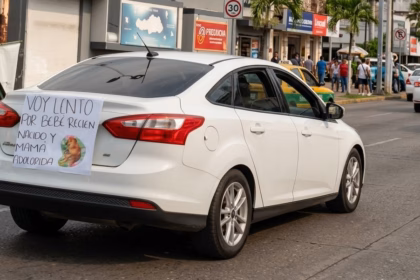 Vista De Tres Cuartos De Un Automóvil Sedán Blanco Circulando Con Un Cartel Pegado En La Cajuela Que Dice &Quot;Voy Lento Por Bebé Recién Nacido Y Mamá Adolorida&Quot;.