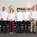Una Fotografía De Grupo Con Seis Funcionarios De Tabasco De Pie Frente A Una Pared Con El Logo Del Gobierno Estatal. De Izquierda A Derecha: Cuatro Hombres Con Guayaberas Blancas Tradicionales Y Pantalones Oscuros O Caqui, Una Mujer Con Blusa Blanca Y Pantalones Color Vino, Y Una Mujer Con Un Conjunto Beige Sin Mangas. Sonríen A La Cámara.