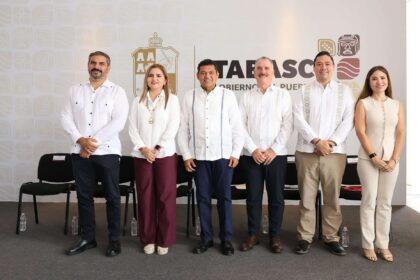 Una Fotografía De Grupo Con Seis Funcionarios De Tabasco De Pie Frente A Una Pared Con El Logo Del Gobierno Estatal. De Izquierda A Derecha: Cuatro Hombres Con Guayaberas Blancas Tradicionales Y Pantalones Oscuros O Caqui, Una Mujer Con Blusa Blanca Y Pantalones Color Vino, Y Una Mujer Con Un Conjunto Beige Sin Mangas. Sonríen A La Cámara.