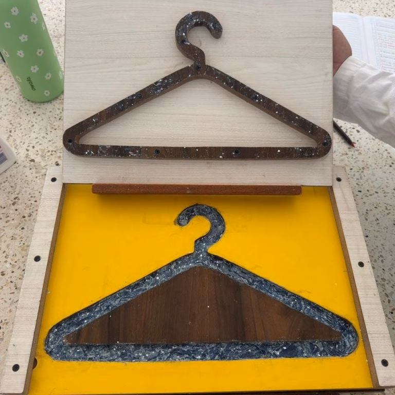 Fotografía De Un Molde Casero Para Fabricar Ganchos De Ropa Ecológicos Utilizando Materiales Reciclados. El Molde Tiene Una Base Amarilla Brillante Con Un Contorno De Resina Azul Oscura Llena De Fragmentos De Mezclilla Triturada Que Delinean La Forma Del Gancho, Y Una Pieza Central De Madera Oscura Rellena El Interior.