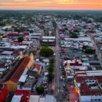 Vista Aérea Panorámica Del Centro Urbano De Cunduacán, Tabasco, Durante El Atardecer, Mostrando Sus Calles, Edificaciones De Techos Rojos Y El Templo Parroquial Iluminado Frente A La Plaza Principal, Rodeados Por La Vegetación Tropical En El Horizonte.