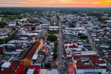 Vista Aérea Panorámica Del Centro Urbano De Cunduacán, Tabasco, Durante El Atardecer, Mostrando Sus Calles, Edificaciones De Techos Rojos Y El Templo Parroquial Iluminado Frente A La Plaza Principal, Rodeados Por La Vegetación Tropical En El Horizonte.