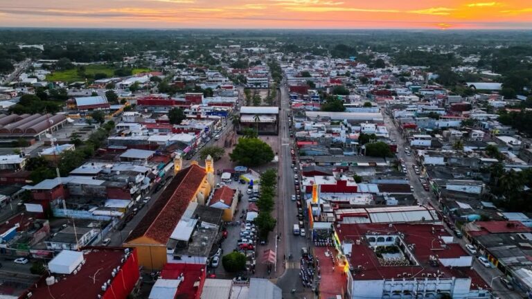 Vista Aérea Panorámica Del Centro Urbano De Cunduacán, Tabasco, Durante El Atardecer, Mostrando Sus Calles, Edificaciones De Techos Rojos Y El Templo Parroquial Iluminado Frente A La Plaza Principal, Rodeados Por La Vegetación Tropical En El Horizonte.