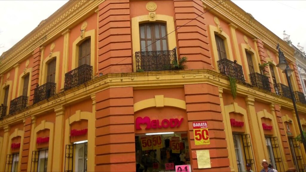 Memoria De Piedra Y Olvido: La Fascinante Historia Del Edificio Del Banco De México En Villahermosa Fachada Esquinera De Un Edificio Histórico De Dos Pisos En Tonos Ocre Y Amarillo, Que En La Planta Baja Alberga El Almacén De Ropa &Quot;Melody&Quot; Con Anuncios De Rebajas.