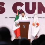 El Gobernador Javier May Rodríguez Dirige Un Mensaje Desde Un Atril De Madera Con El Escudo De Tabasco, Flanqueado Por La Bandera De México Y La Bandera Estatal, Frente A Una Audiencia En Un Recinto Oficial.