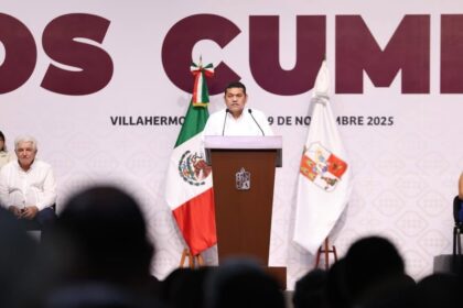 El Gobernador Javier May Rodríguez Dirige Un Mensaje Desde Un Atril De Madera Con El Escudo De Tabasco, Flanqueado Por La Bandera De México Y La Bandera Estatal, Frente A Una Audiencia En Un Recinto Oficial.
