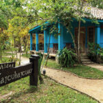 Cabaña De Color Azul Rodeada De Vegetación Tropical En Tacotalpa, Con Un Letrero De Madera En Primer Plano Que Indica El Acceso Al Complejo Turístico.