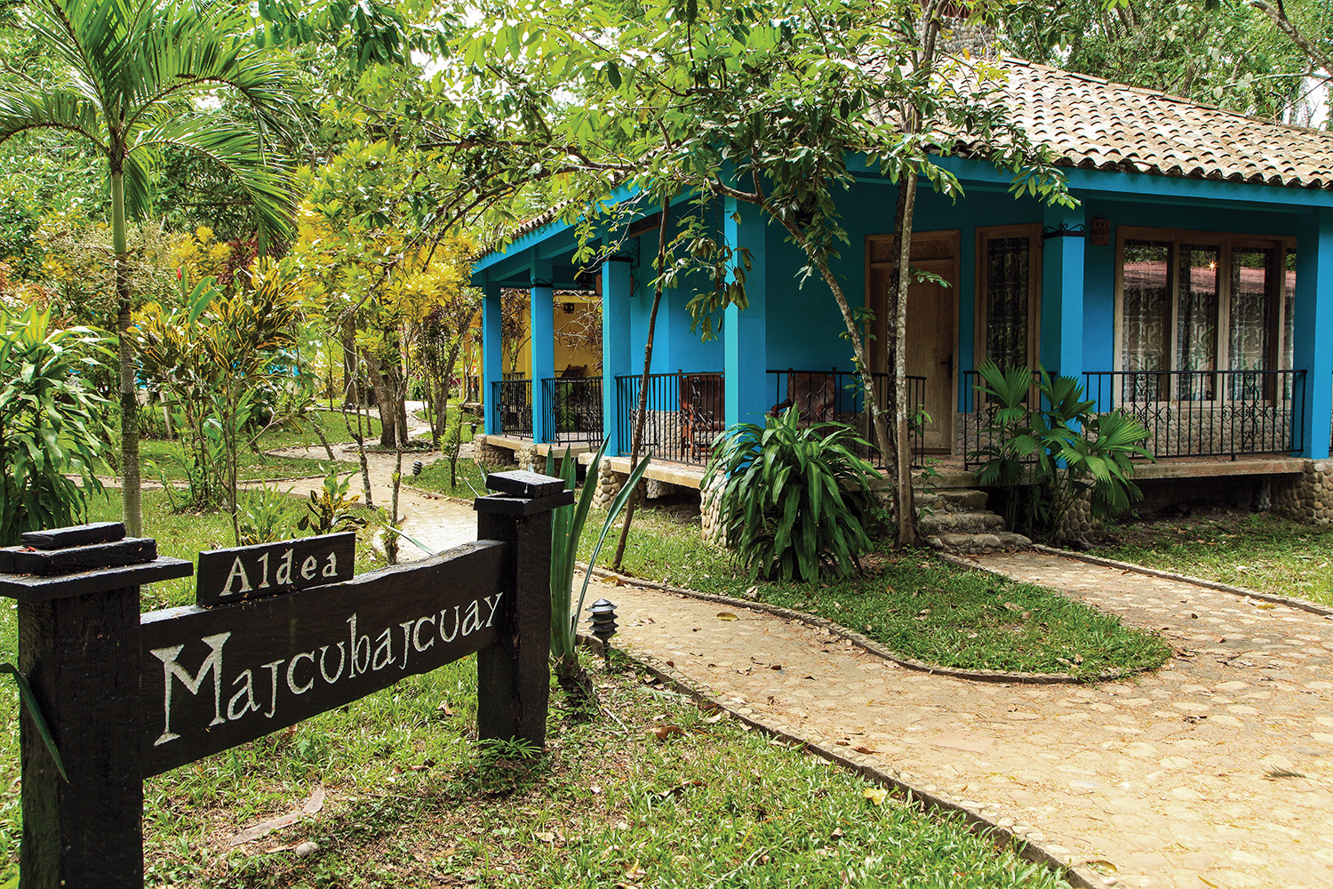Descubre Tacotalpa: Tu Próxima Escapada De Fin De Semana En Cabañas Junto Al Río Cabaña De Color Azul Rodeada De Vegetación Tropical En Tacotalpa, Con Un Letrero De Madera En Primer Plano Que Indica El Acceso Al Complejo Turístico.