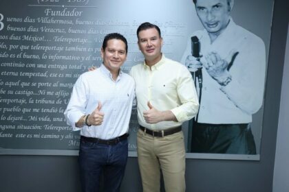 Jesús Abraham Cano González, Conocido Popularmente Como “Chelo Cano”, Y Emmanuel Sibilla Oropesa Sonriendo Y Mostrando El Pulgar Hacia Arriba En Señal De Aprobación, Parados Frente A Un Mural Con Texto Y La Imagen De Don Jesús Antonio Sibilla Zurita En Las Instalaciones De La Xevt.