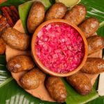Plato De Barro Con Butifarras De Jalpa Dispuestas Alrededor De Un Recipiente Central Con Cebolla Morada Picada, Acompañado De Plátanos Fritos Y Tortillas Sobre Hojas De Plátano.