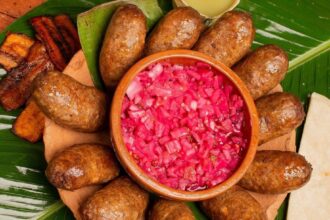 Plato De Barro Con Butifarras De Jalpa Dispuestas Alrededor De Un Recipiente Central Con Cebolla Morada Picada, Acompañado De Plátanos Fritos Y Tortillas Sobre Hojas De Plátano.
